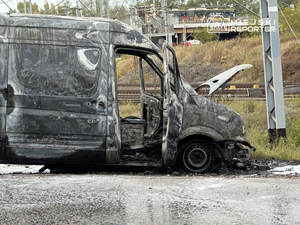 Spalony furgon stojący na poboczu drogi, z widocznymi uszkodzeniami karoserii i zniszczonym wnętrzem.