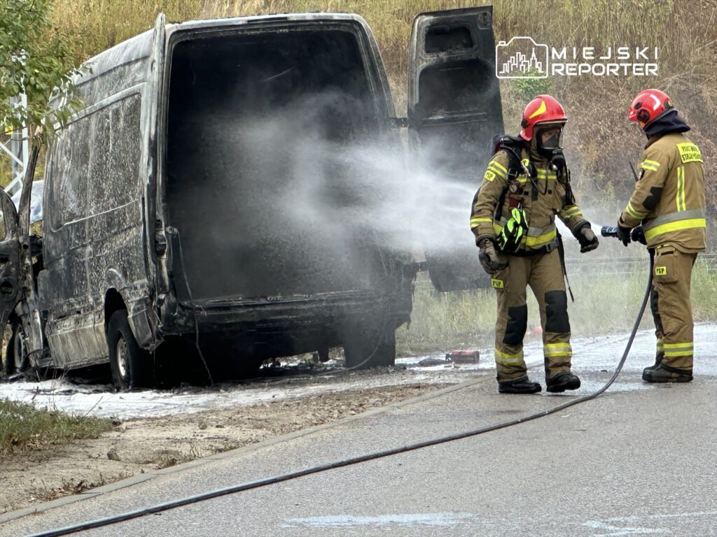 Strażacy w mundurach gaśniczych gaszą palący się pojazd dostawczy przy drodze, obok wydobywa się dym.