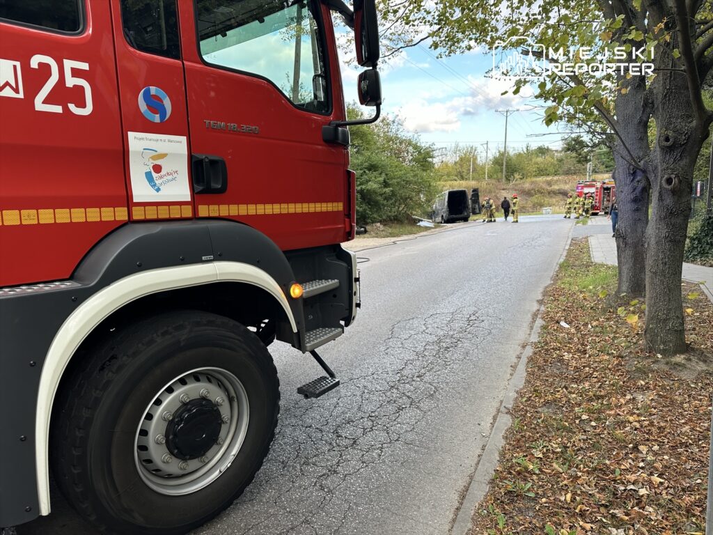 Czerwony wóz strażacki stoi na drodze, obok grupy strażaków pracujących przy przewróconym pojeździe.