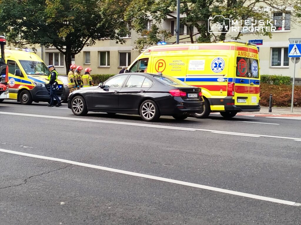 Karetki pogotowia oraz strażacy rozmawiają z policjantem na ulicy obok czarnego samochodu osobowego.