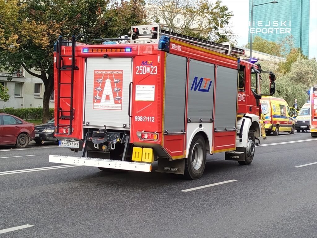 Czerwony wóz strażacki z włączonymi sygnalizatorami przejeżdża ulicą, obok zaparkowanych samochodów i ambulansów.