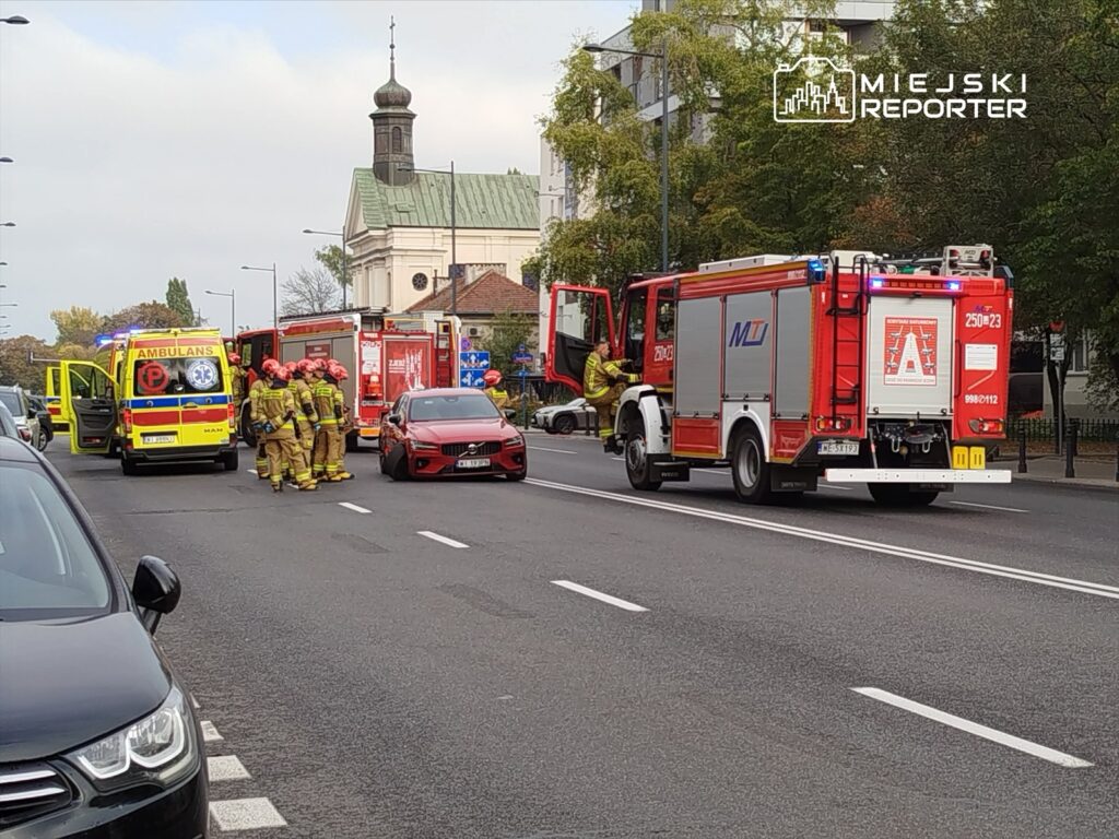 Grupa strażaków w kombinezonach ratunkowych omawia sytuację obok wozu strażackiego na ulicy, obok stoi karetka.
