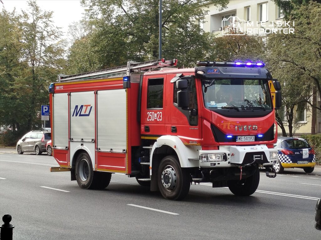 Wozidło strażackie marki Iveco z niebieskimi światłami przejeżdża ulicą w otoczeniu drzew i zaparkowanych samochodów.