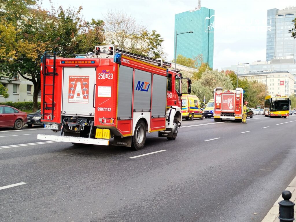 Wozy strażackie przejeżdżające obok zaparkowanych samochodów w miejskim otoczeniu, z widocznymi pojazdami służb ratunkowych.