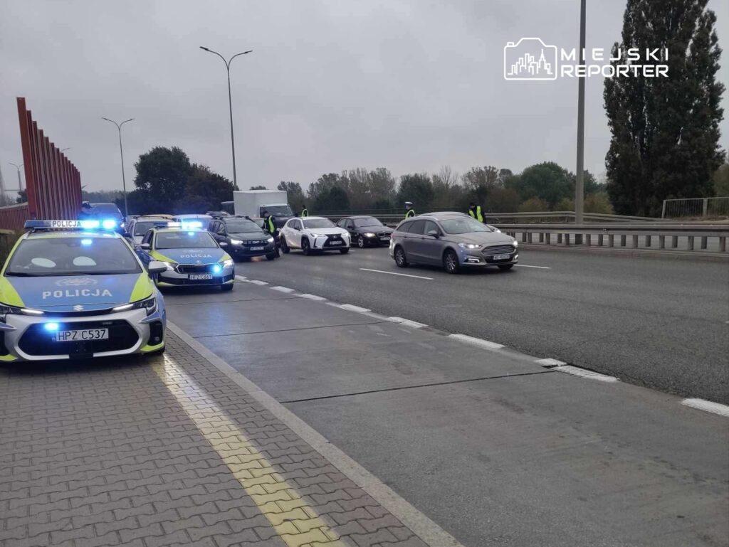 Policyjne radiowozy zatrzymują ruch na drodze, a funkcjonariusze kontrolują pojazdy w deszczową pogodę.
