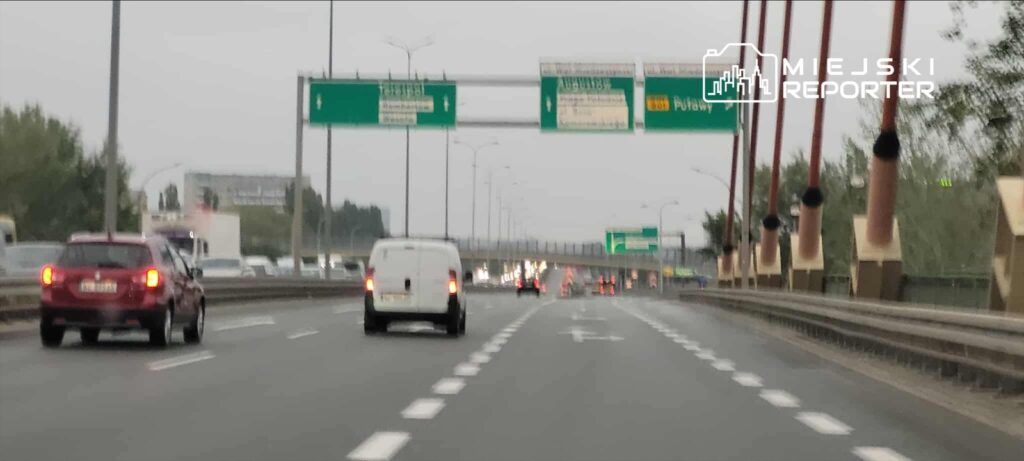 Ruch samochodowy na dwupasmowej drodze, z widocznymi znakami kierunkowymi oraz szarym niebem.