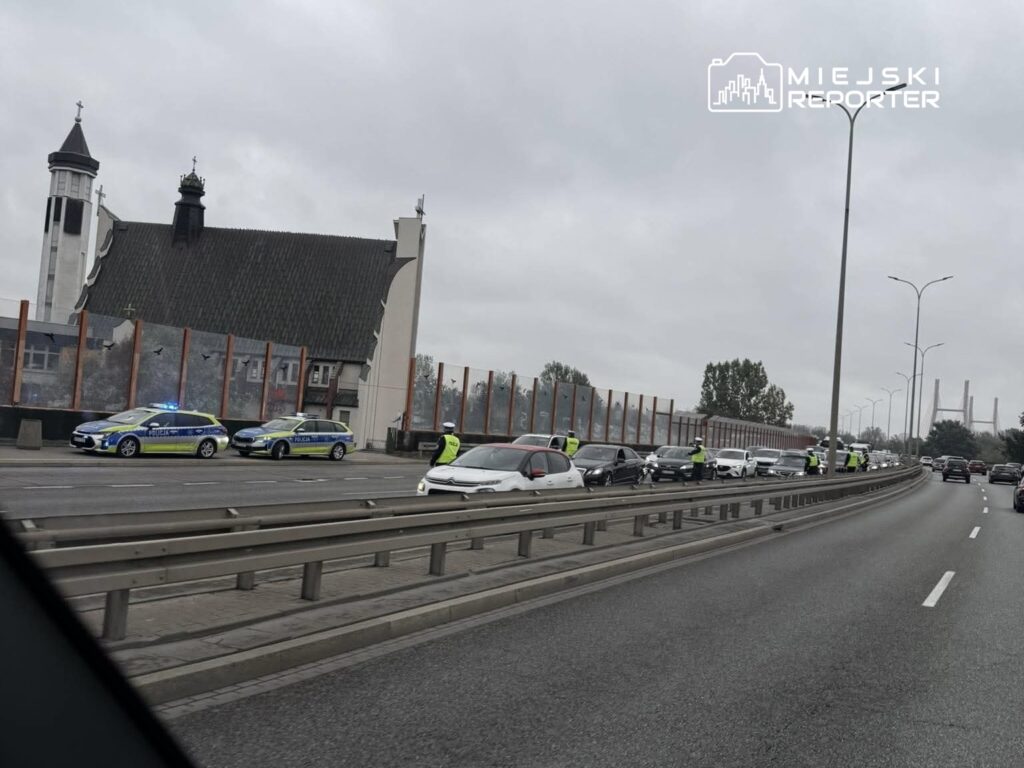 Policjanci kontrolują ruch drogowy na moście, obok nowoczesnego kościoła, w dzień pochmurny.