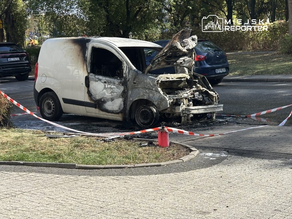 Spalony biały furgon z otwartą maską, otoczony taśmą ostrzegawczą, na miejskiej ulicy.
