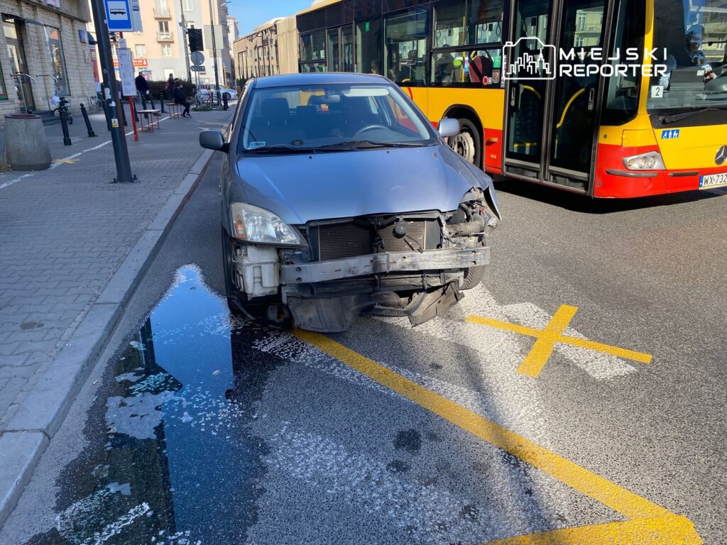 Uszkodzony samochód zaparkowany na ulicy obok przystanku autobusowego, w tle przejeżdżający autobus.