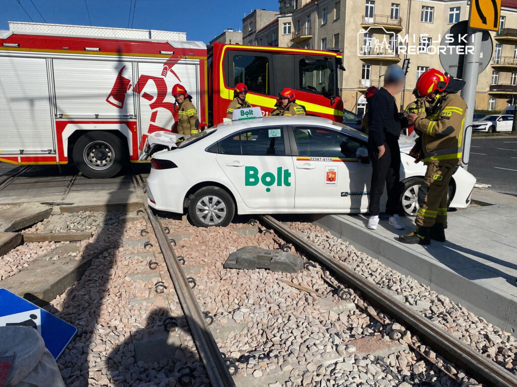 Kierowca w czarnej bluzie rozmawia z ratownikami obok białego samochodu Bolt, który utknął na torach tramwajowych.