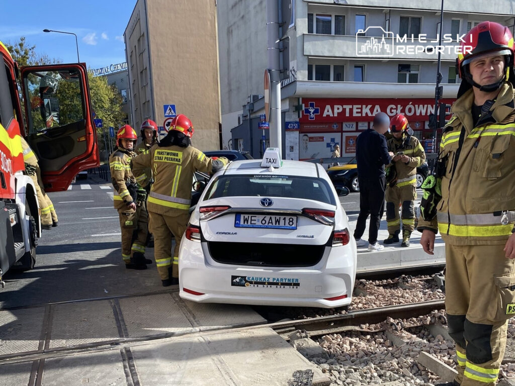 Grupa strażaków w mundurach bada uszkodzone białe taxi stojące na torach tramwajowych w mieście.
