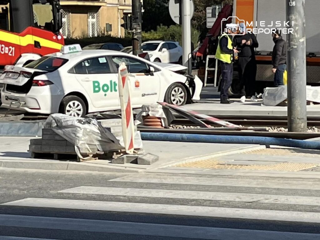 Biały samochód osobowy z logo Bolta stoi uszkodzony na skrzyżowaniu, obok grupy osób w odzieży roboczej oraz straży pożarnej.