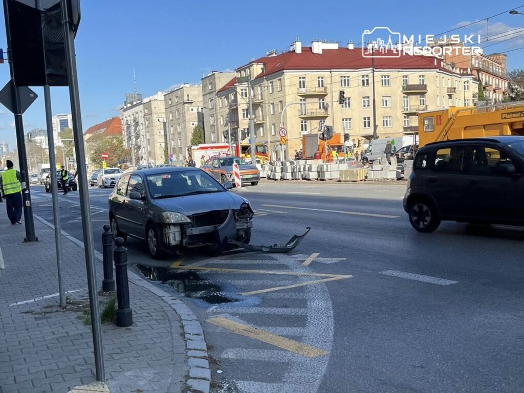 Uszkodzony samochód osobowy stoi na pasie drogowym obok robotników w kamizelkach odblaskowych, w tle prace budowlane.