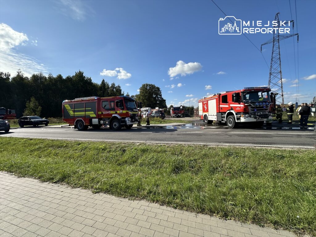 Czerwony wóz strażacki stojący na drodze obok akcji ratunkowej, w tle zalesiony teren i niebo z chmurami.