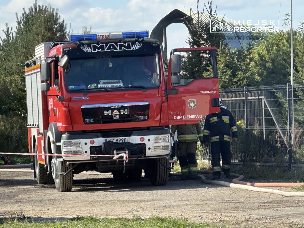 Czerwony wóz strażacki MAN z otwartą kabiną, obok strażacy w mundurach przygotowujący się do akcji.
