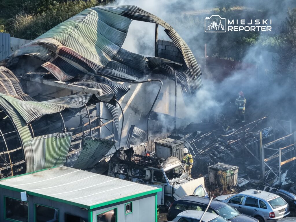 Zniszczony budynek z metalowym dachem, nad którym unosi się dym, w otoczeniu spalonego wraku pojazdu i strażaków gaszących ogień.