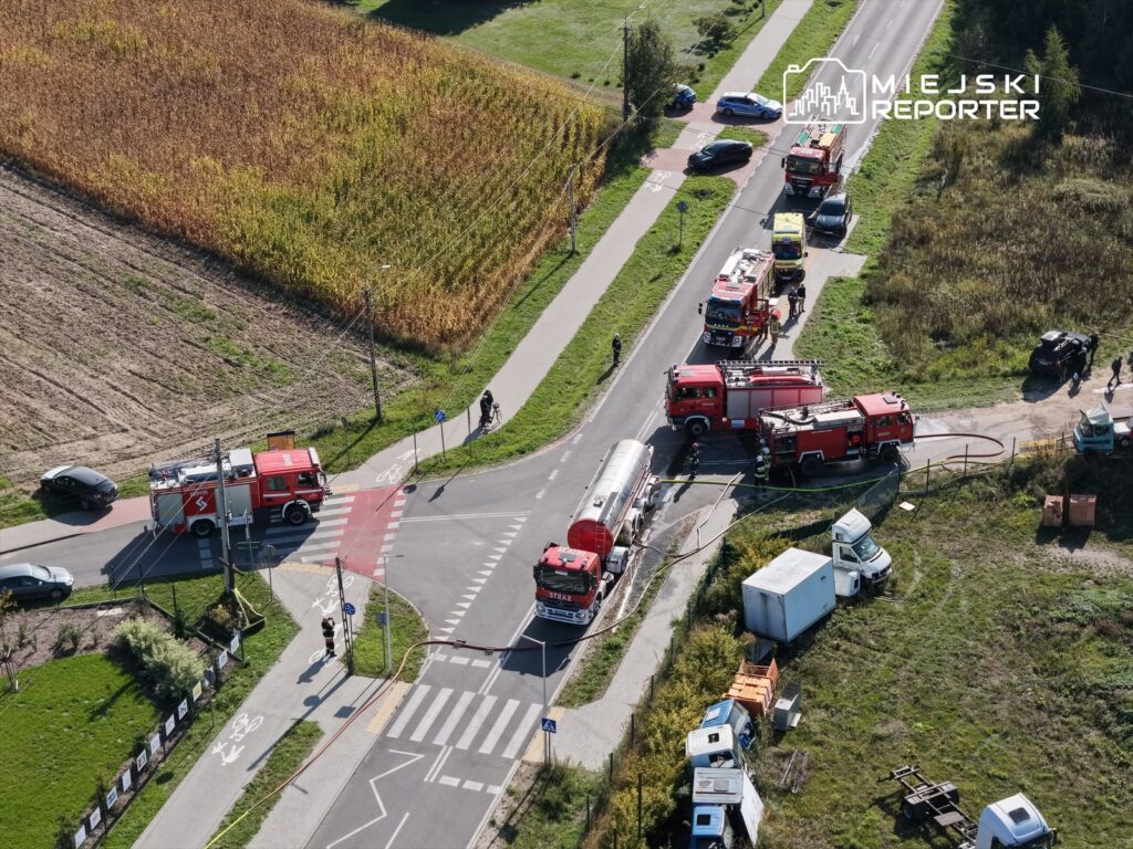 Na skrzyżowaniu dróg stoją wozy strażackie, a obok pracują strażacy. W tle pola i zarośla.