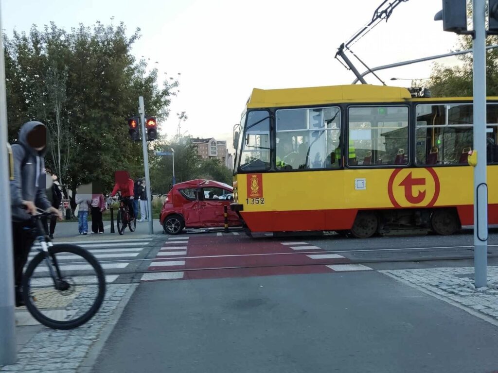 Zderzenie tramwaju z czerwoną osobówką na skrzyżowaniu, obok grupy przechodniów i rowerzysty.