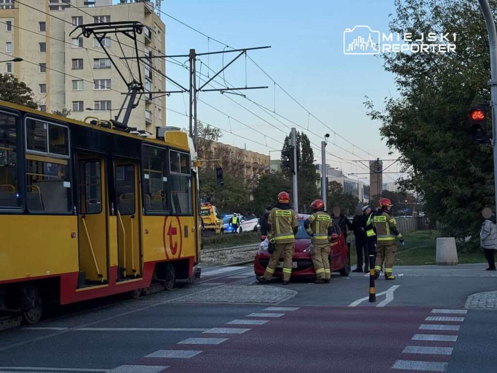 Czerwony samochód zatrzymany na skrzyżowaniu, obok trzech strażaków w hełmach, którzy udzielają pomocy poszkodowanej osobie.