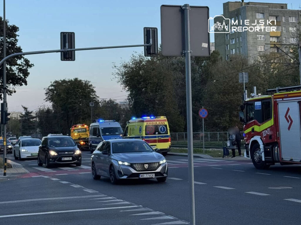 Samochody osobowe zatrzymane na drodze, w tle karetka i wóz strażacki z włączonymi sygnałami świetlnymi.