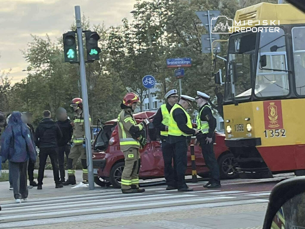 Grupa strażaków i policjantów rozmawia obok uszkodzonego czerwonego samochodu na skrzyżowaniu z tramwajem.