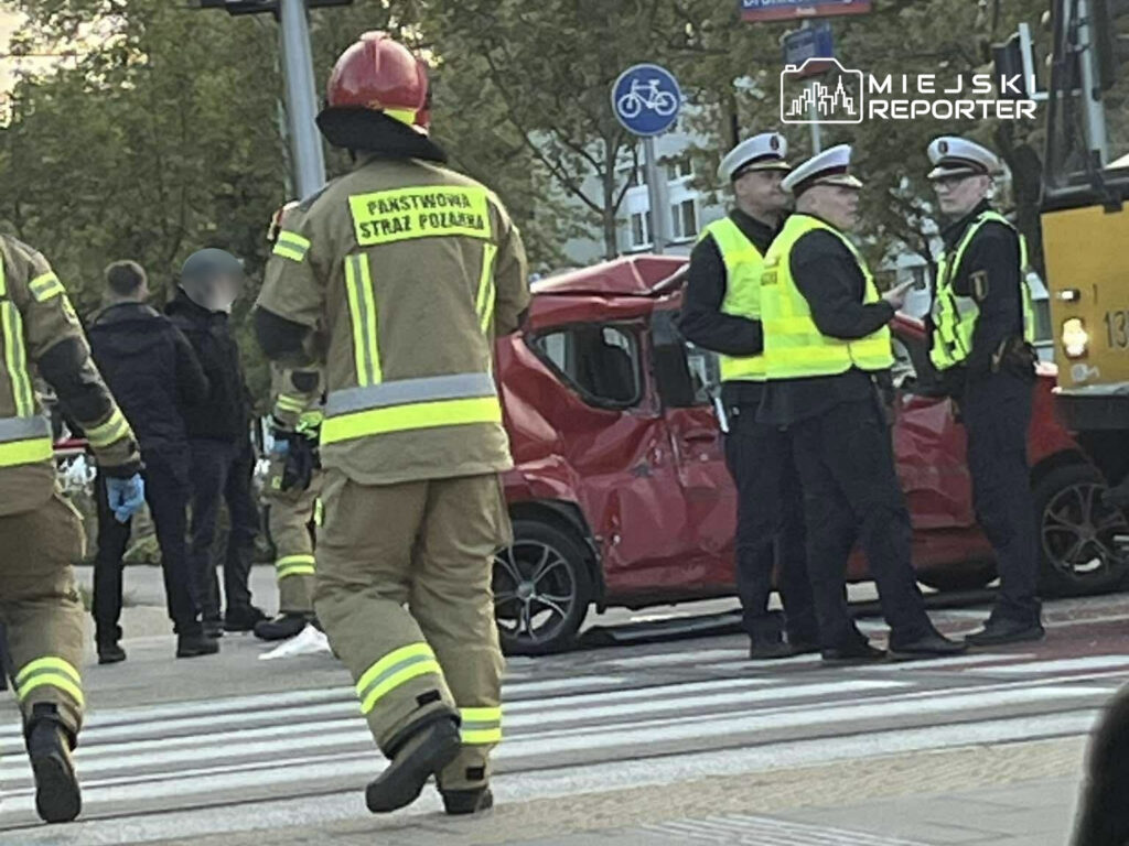 Grupa strażaków i policjantów rozmawia obok uszkodzonego czerwonego samochodu na przejściu dla pieszych.