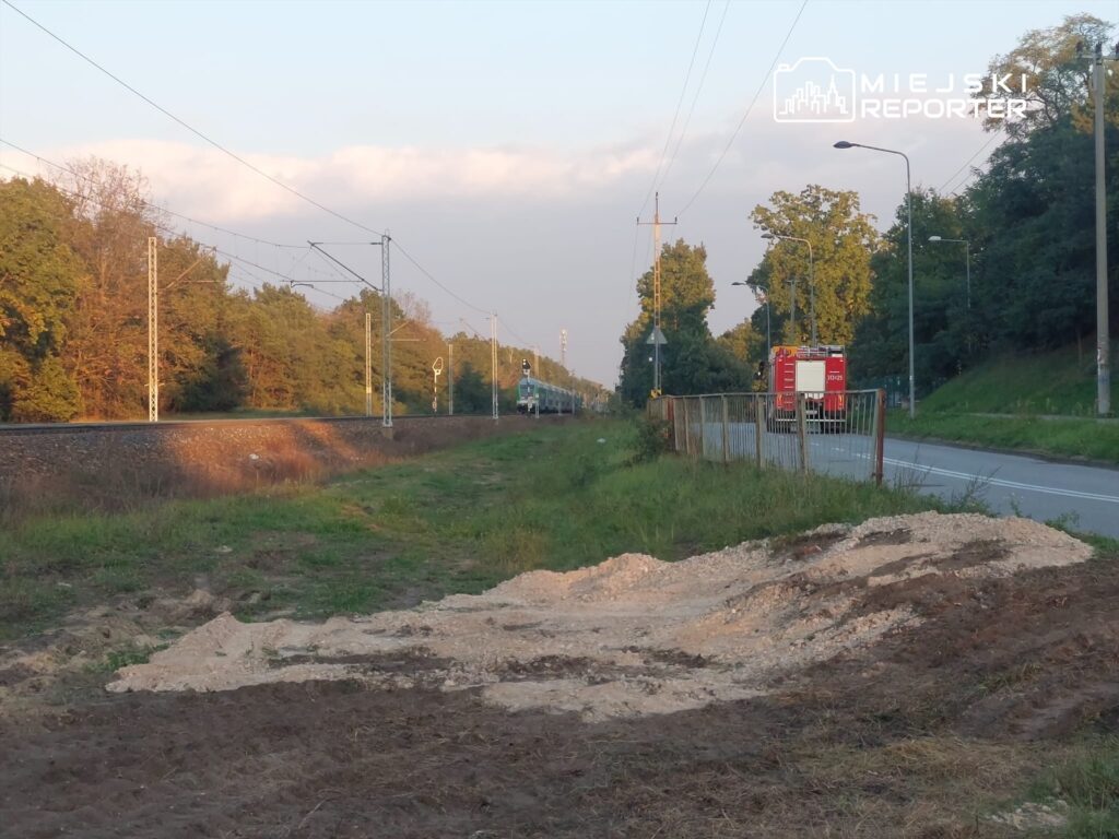 Na poboczu drogi leży wykopany grunt, obok torów kolejowych, w tle widać pociąg i strażacki wóz.