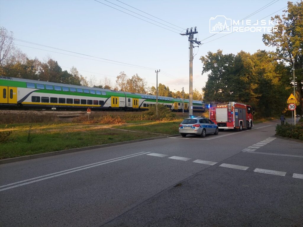 Pociąg osobowy w zielono-żółtej kolorystyce przejeżdża obok drogi, obok której stoją wóz strażacki i radiowóz policyjny.