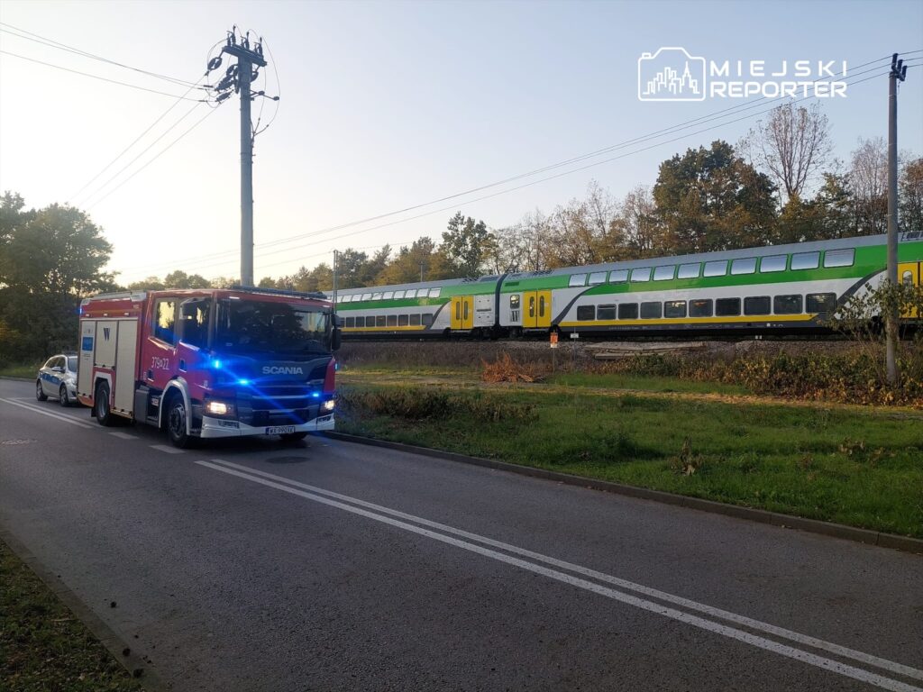 Wozidło strażackie zatrzymane obok drogi, podczas gdy pociąg pasażerski przejeżdża w tle.