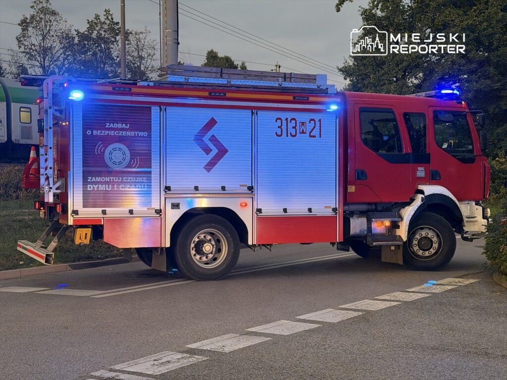 Czerwony wóz strażacki z włączonymi sygnalizatorami świetlnymi stoi na poboczu drogi, obok torów kolejowych. Widać napis „ZADBaj O BEZPIECZEŃSTWO ZAMONTUJ CZUJKE DYMU I GAZU”.