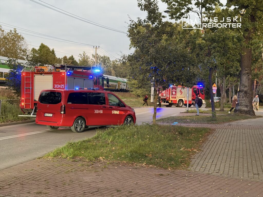 Czerwony wóz strażacki i ambulans zatrzymane na ulicy, w tle pociąg przejeżdżający obok. Przechodnie idą chodnikiem.