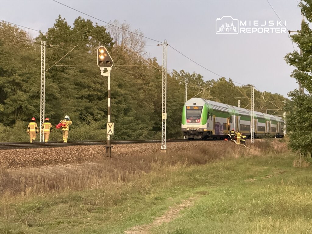 Trzech strażaków w żółtych mundurach idzie wzdłuż torów kolejowych obok pociągu pasażerskiego.