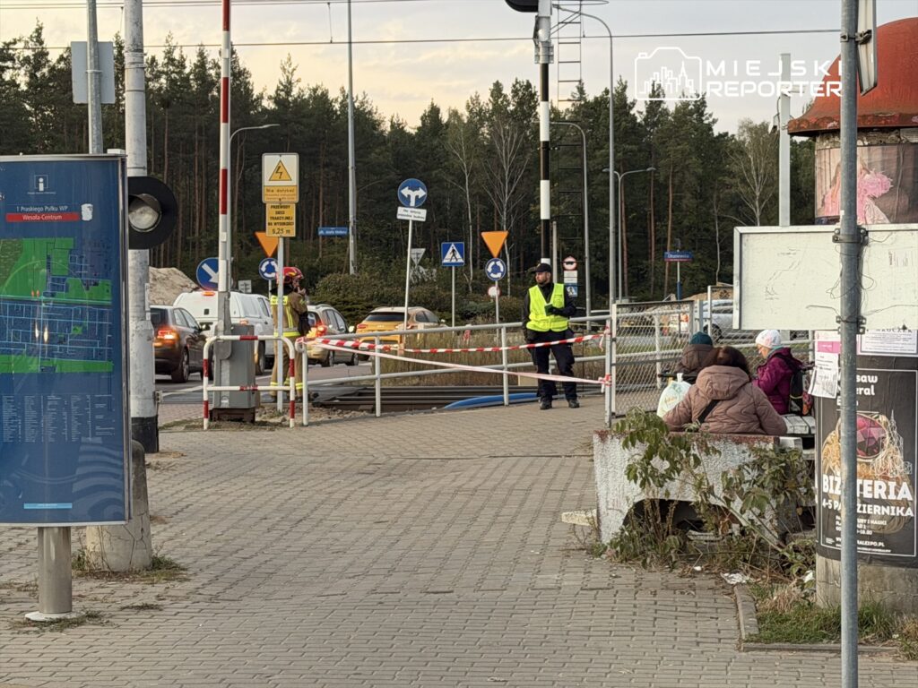 Ruch drogowy zatrzymany przez strażnika w kamizelce odblaskowej przy przejeździe kolejowym, obok czekających pasażerów.