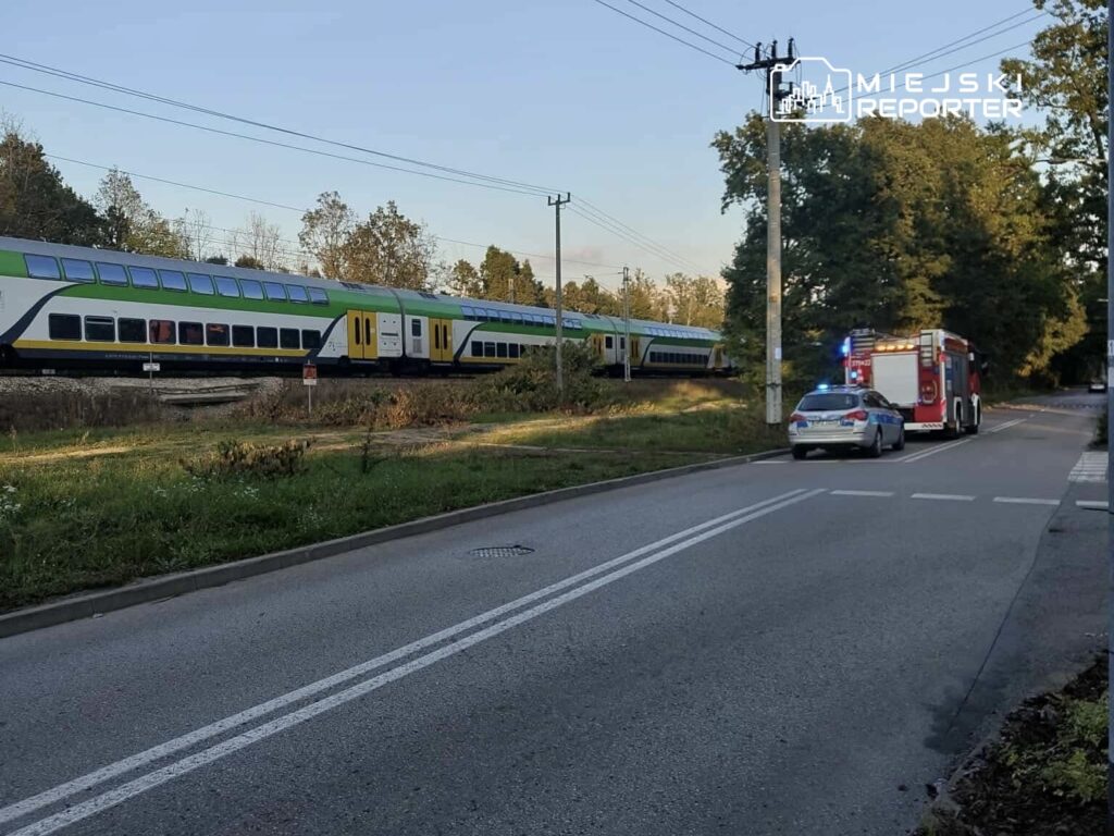Pociąg osobowy przejeżdża obok drogi, obok której stoją samochód policyjny i wóz strażacki.