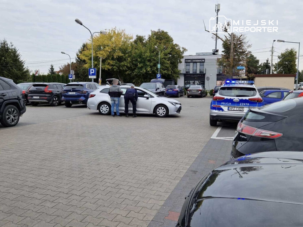 Mężczyzna w dżinsach i koszuli rozmawia z policjantem obok białego samochodu na parkingu.