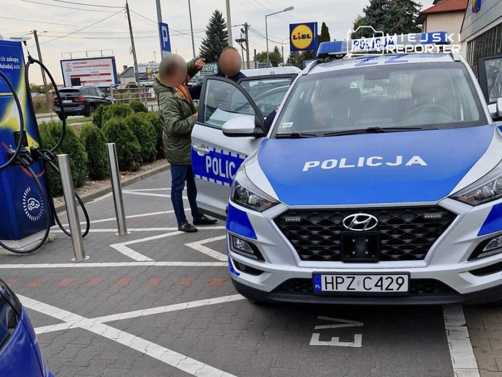 Mężczyzna w zielonej kurtce rozmawia z policjantem przy radiowozie na stacji paliw.