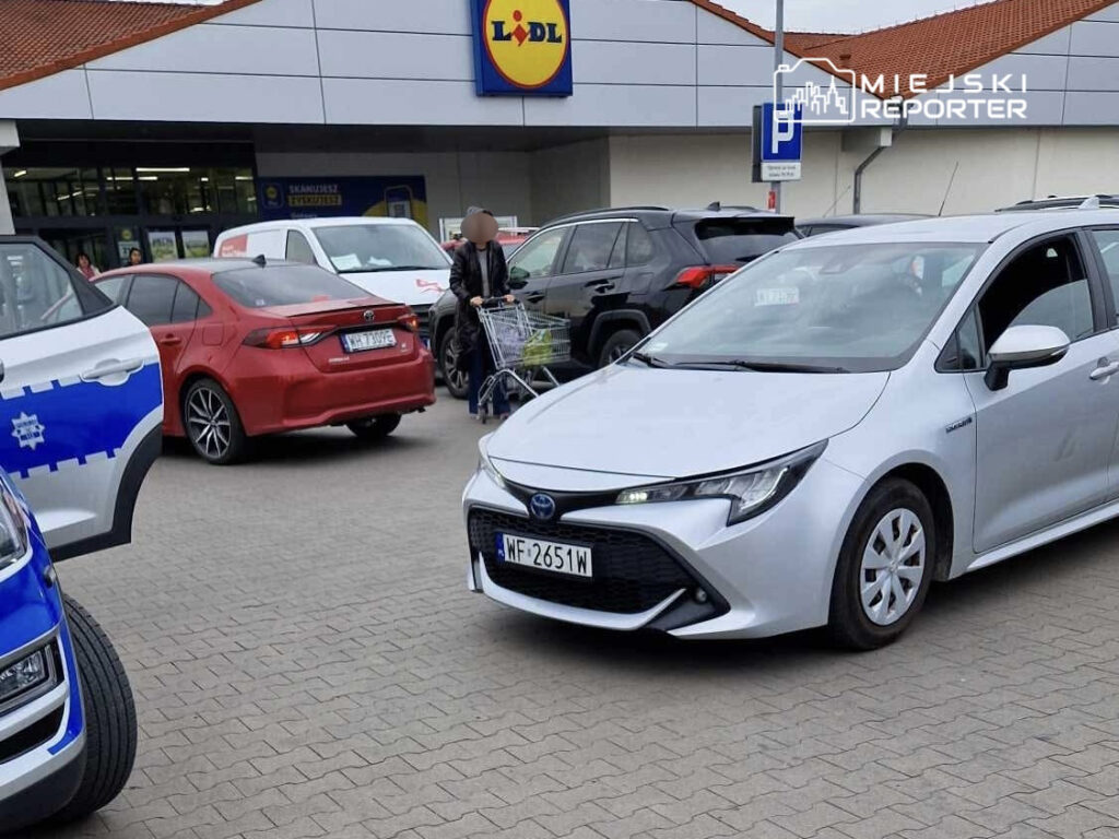 Na parkingu przed sklepem Lidl zaparkowane są różne samochody, w tym srebrna Toyota oraz czerwona limuzyna. Klientka z wózkiem na zakupy zmierza w kierunku wejścia do sklepu.