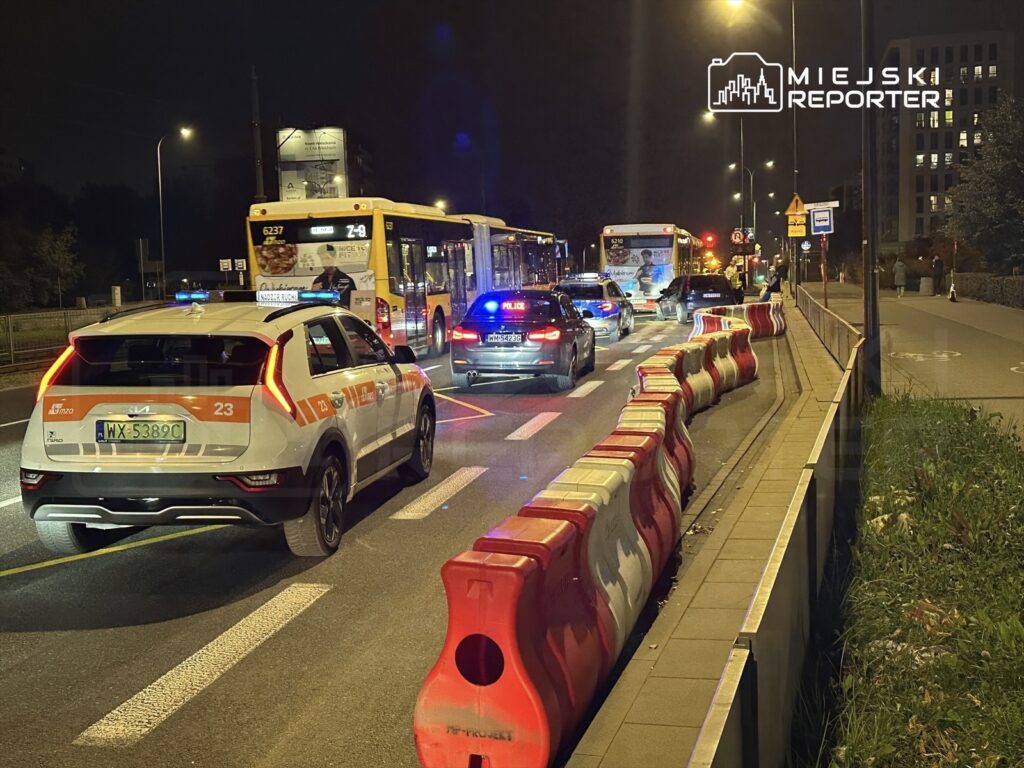 Policyjny radiowóz z włączonymi sygnałami świetlnymi zatrzymuje ruch na ulicy, obok stoją autobusy.
