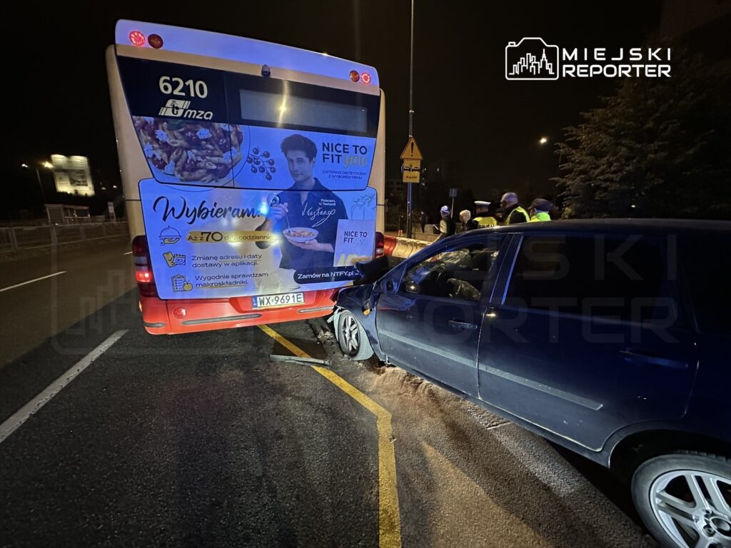 Zderzenie autobusu miejskiego z osobowym samochodem na ulicy w nocy, w tle widoczni świadkowie zdarzenia.