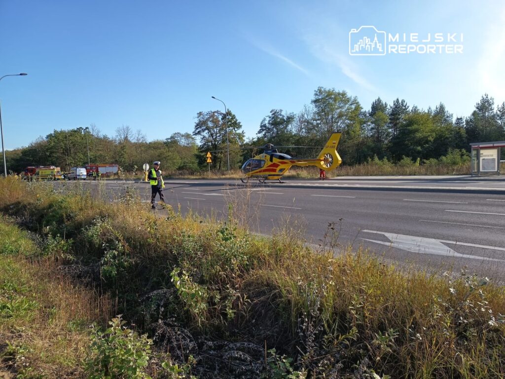 Policjant w kamizelce odblaskowej kieruje ruchem obok lądowiska helikoptera ratunkowego na drodze.