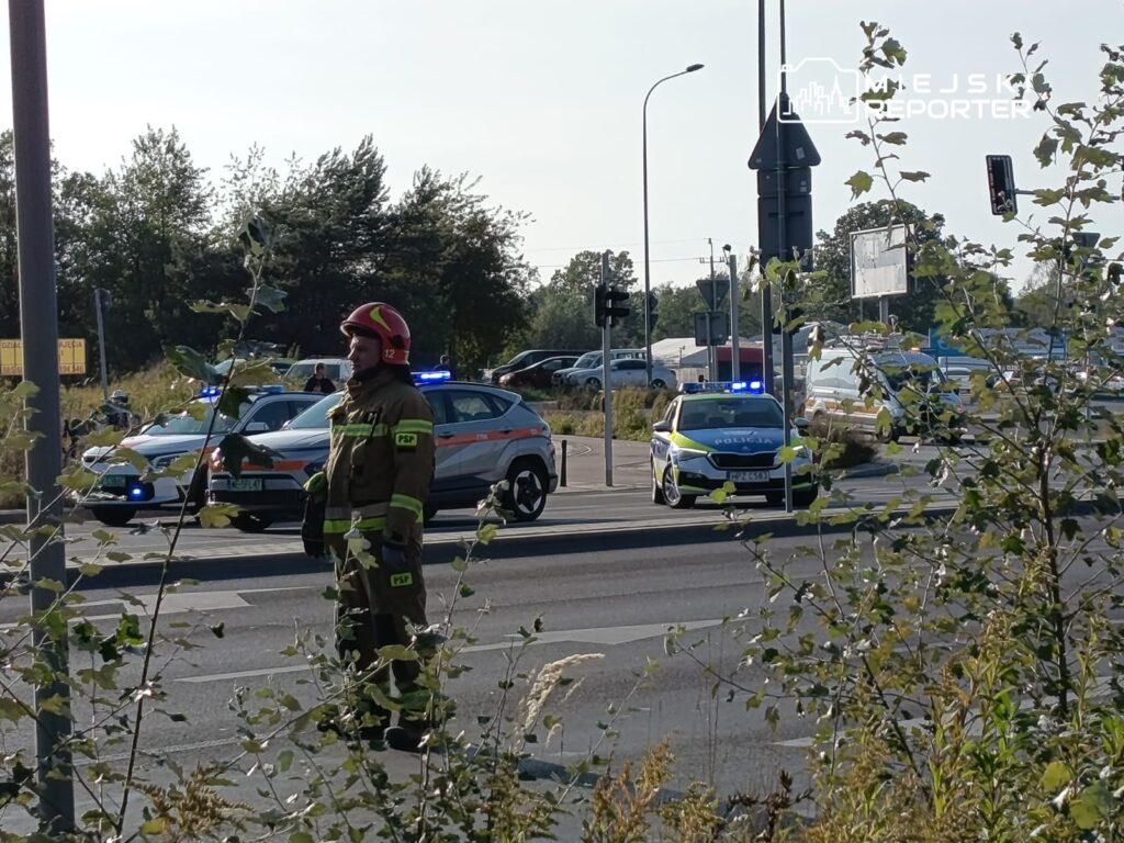 Strażak w hełmie kieruje ruchem na skrzyżowaniu, obok policyjne radiowozy z włączonymi sygnałami świetlnymi.