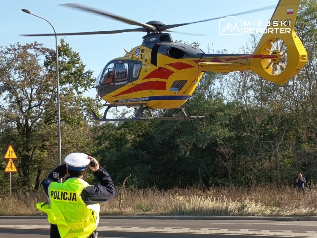 Policjant w żółtej kamizelce fotografuje przelatujący śmigłowiec ratunkowy nad drogą w otoczeniu drzew.