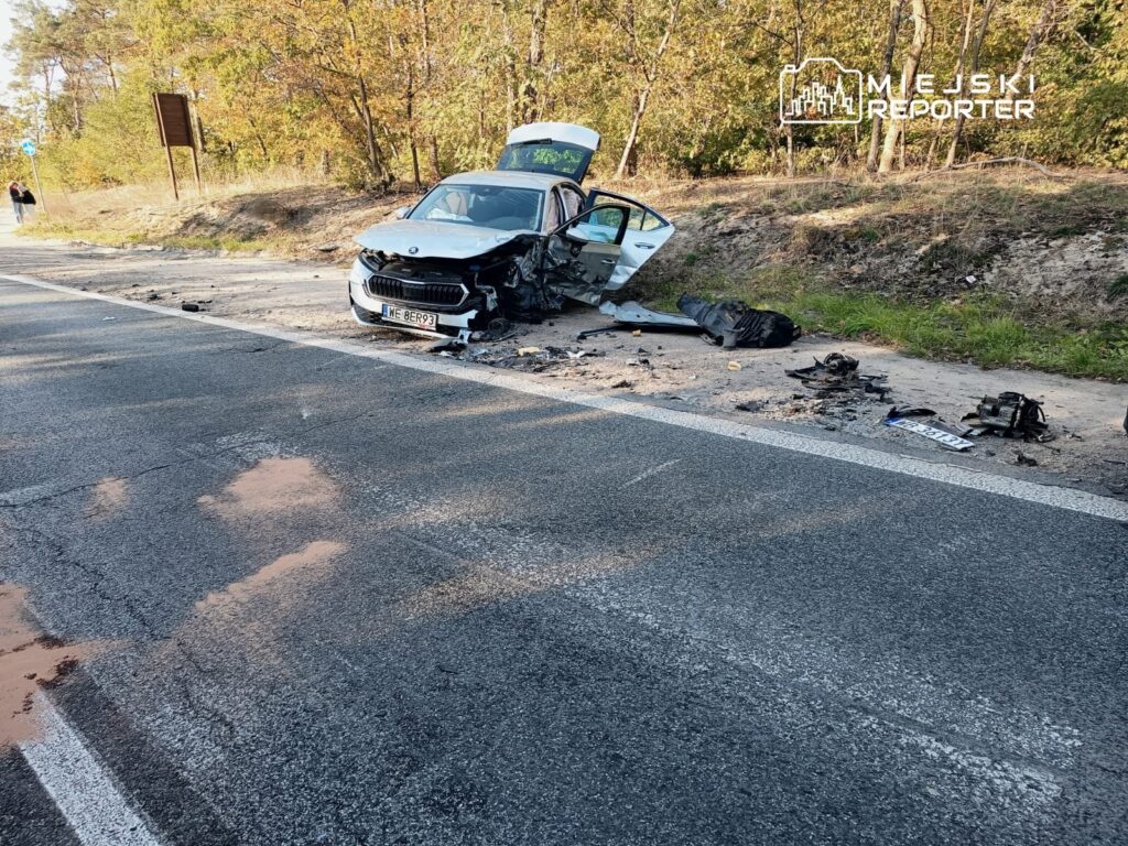 Zniszczony samochód osobowy stoi na poboczu drogi, obok leżą fragmenty wraku i plamy oleju. W tle widoczna zieleń drzew.