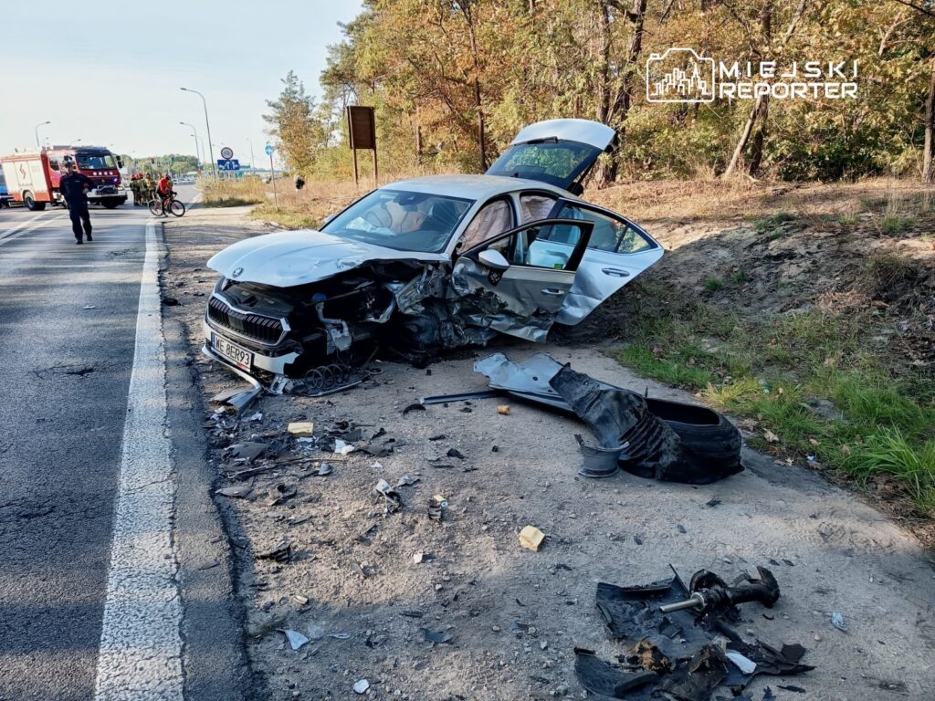 Uszkodzony samochód osobowy stoi na poboczu drogi, obok leżą fragmenty karoserii i opon. W tle widać straż pożarną oraz rowerzystów.