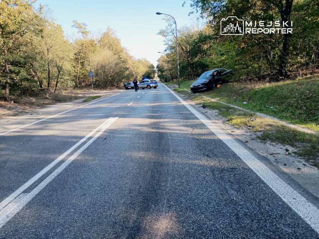 Na drodze leżącej wśród drzew stoi czarne auto, a obok znajdują się policjanci prowadzący czynności.