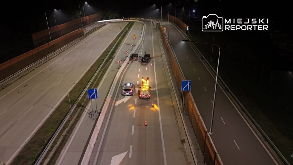 Roboty drogowe prowadzone na nocnej jezdni, z oznakowaniem i pojazdem zabezpieczającym w akcji.