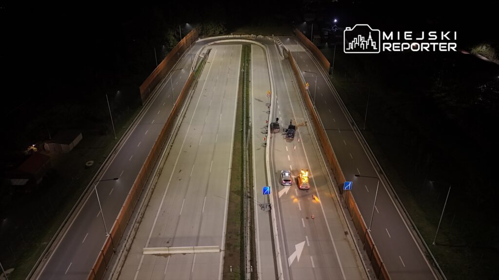 Na drodze nocą stoją dwa samochody służbowe, a w tle widoczne są bariery i lampy oświetlające jezdnię.