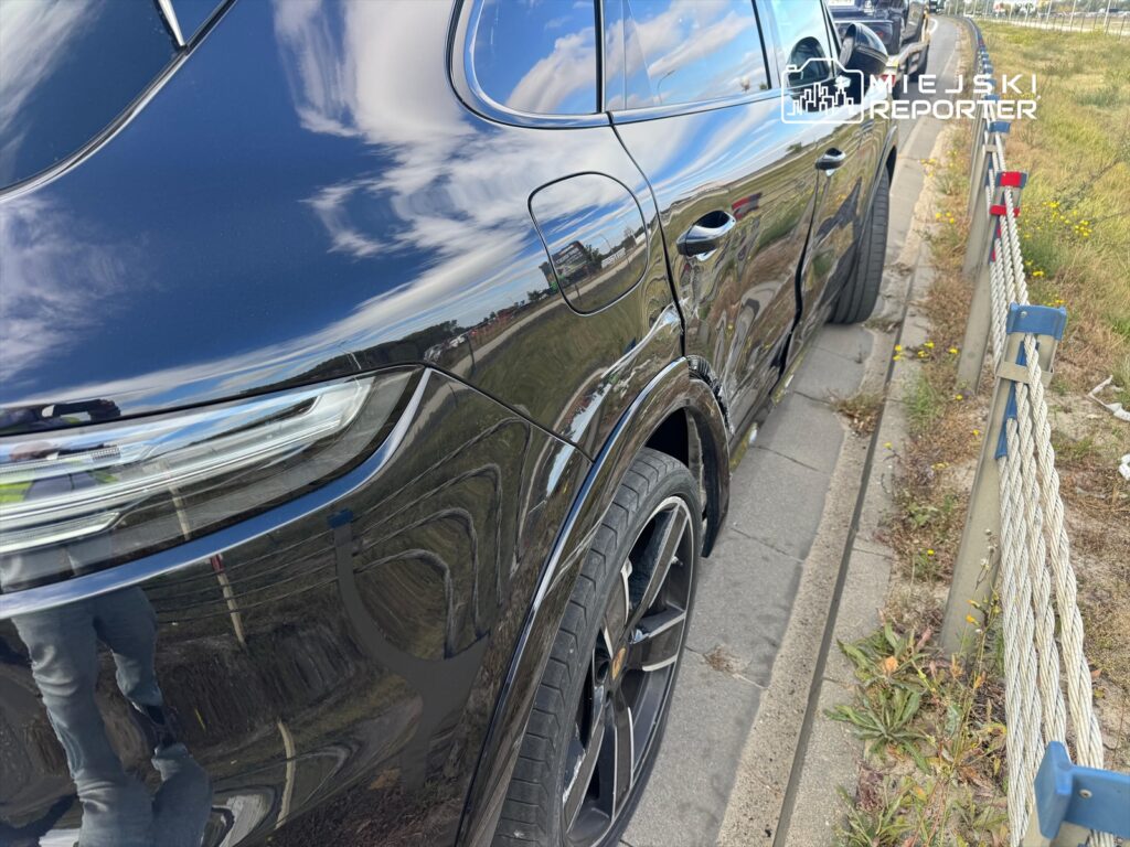 Czarny SUV z widoczną rysą na nadwoziu zaparkowany przy chodniku w otoczeniu trawy i ogrodzenia.