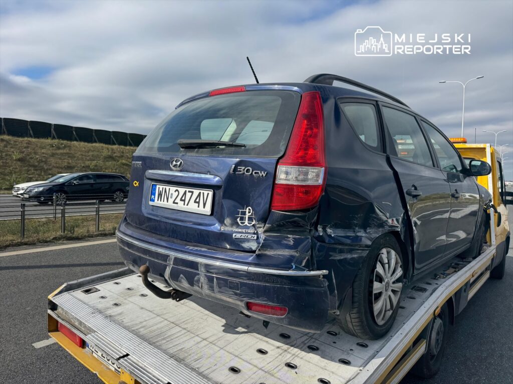 Uszkodzony niebieski samochód osobowy na lawecie, stojący na poboczu ruchliwej drogi.
