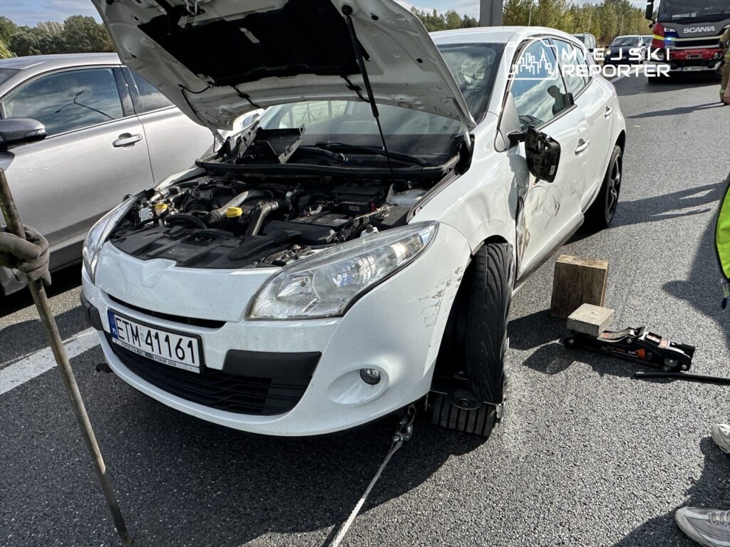 Biały samochód osobowy z otwartą maską, stojący na drodze, obok widoczne narzędzia do naprawy.